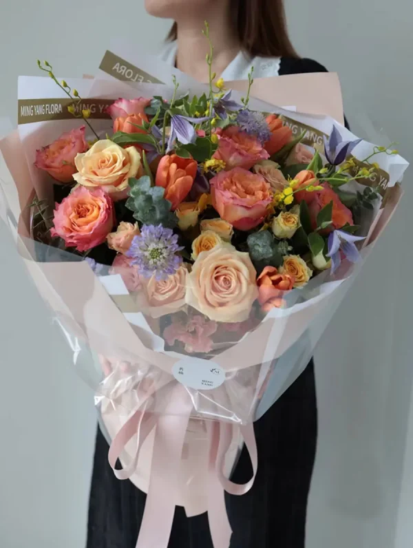 Real Photo - Florist Holding Sunset Glow Mixed Bouquet Real-life photo of a florist holding the massive Sunset Glow mixed bouquet of peach roses, vibrant tulips, and purple accents. Demonstrates the actual size and lush texture for China flower delivery.